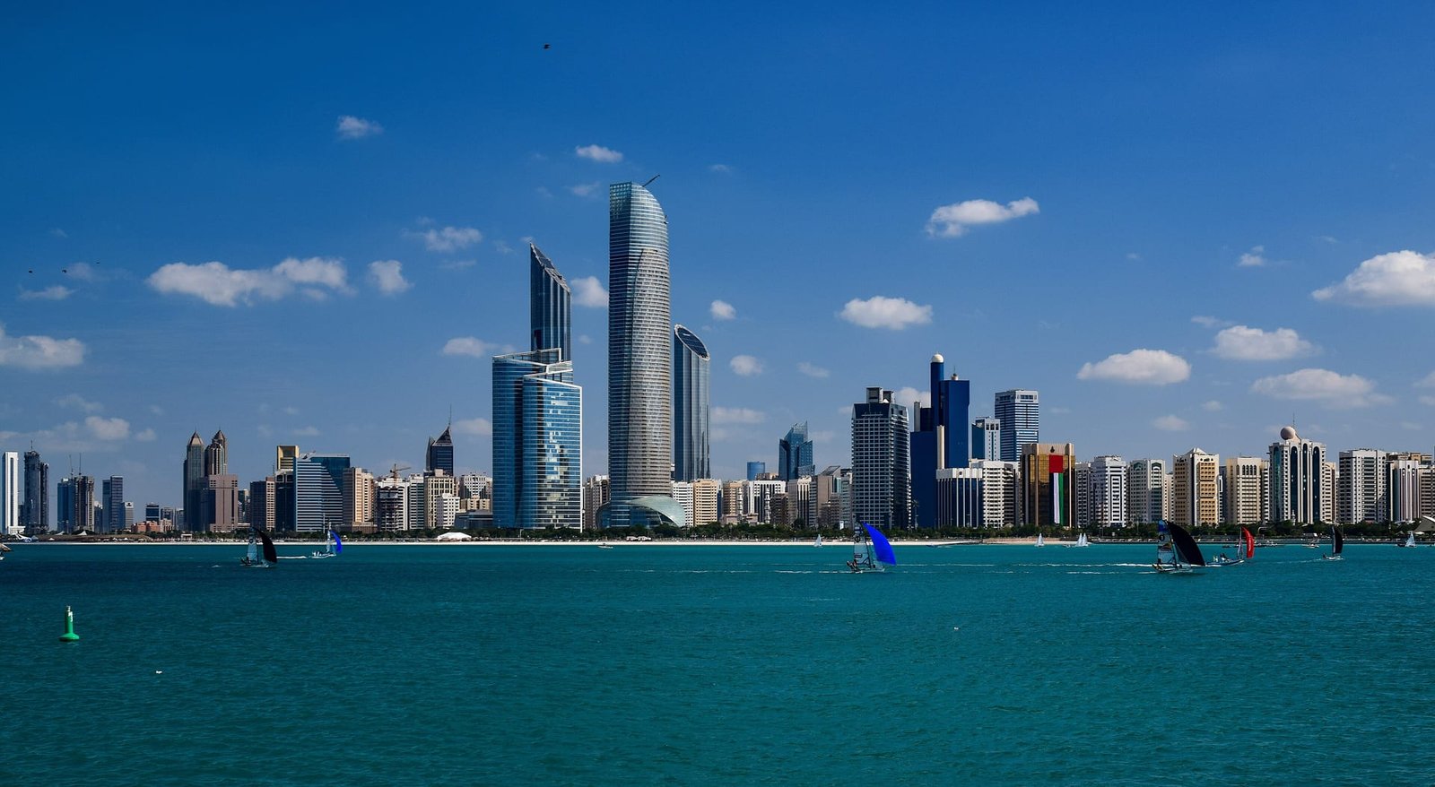 A cluster of curved-glass towers on the Abu Dhabi Corniche — mirror-finish facades, sweeping sea-front approaches and floodlit crowns after dark.